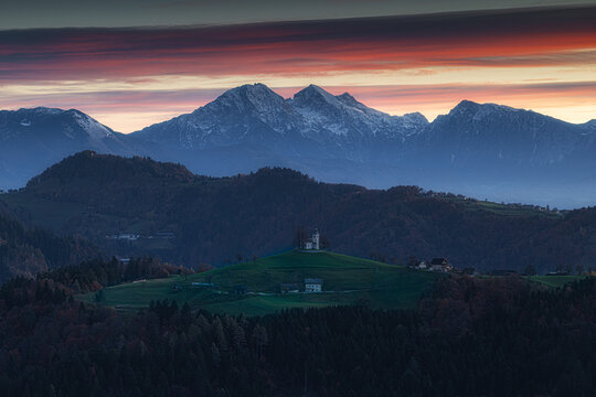 Aerial view of a solitary church atop a verdant hill against a backdrop of snow-dusted mountains beneath a fiery sunset sky, Spodnja Lua, Slovenia.