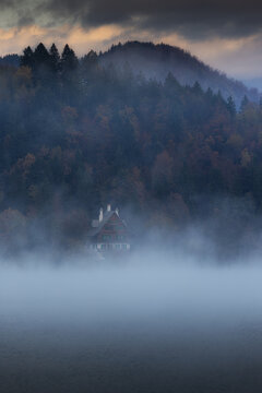 Aerial view of a serene house emerges from the misty embrace, nestled against a backdrop of the dark forest, Spodnja Lua, kofja Loka, Slovenia.