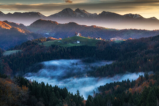 Aerial view of the church atop the hill surrounded by mist and forest with mountains in the background, Spodnja Lua, kofja Loka, Slovenia.