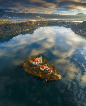 Aerial view of the church on Bled Island reflected in the still lake waters, surrounded by autumnal colors under a dramatic sky, Spodnja Lua, kofja Loka, Slovenia.