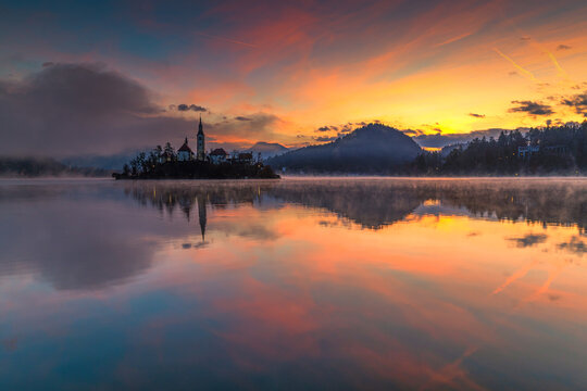 Aerial view of the church steeple piercing the golden dawn light over the still waters of the lake, reflecting the sky's pastel hues, Spodnja Lua, kofja Loka, Slovenia.