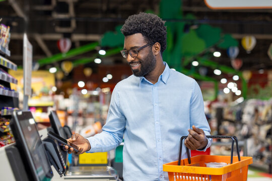 Young smiling african american man paying contactless with smartphone at a self-checkout terminal in modern supermarket, holding a shopping basket with groceries