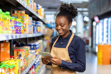 Smiling young african american woman wearing an apron and a dotted shirt, viewing content on a digital tablet while checking products on shelves in a supermarket aisle