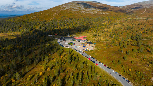 Aerial view of a building complex nestled amidst a vibrant landscape of dark green forests and golden tundra under a vast sky, Enontekio, Lapland, Finland.