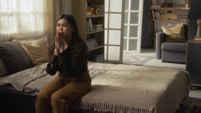 Caucasian gen Z woman with long hair folding hands together and whispering prayer while sitting on bed in her apartment
