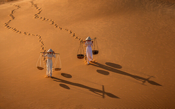 Vietnamese ladies in traditional ao dai white dresses and conical hats traverse the undulating golden dunes. They carry straw baskets, their shadows stretching, Mui Ne, Phan Thiet, Vietnam.