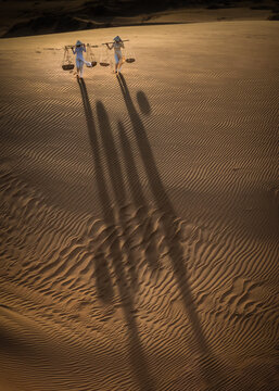Aerial view of two Vietnamese ladies traversing the golden dunes, their elongated shadows stretching across the rippled sands, Mui Ne, Phan Thiet, Vietnam.