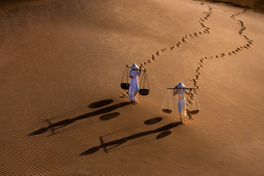 Two Vietnamese women in traditional ao dai dresses traverse the undulating dunes, leaving a trail of footprints in the golden sand. They carry straw baskets. Long shadows. Mui Ne, Phan Thiet, Vietnam.