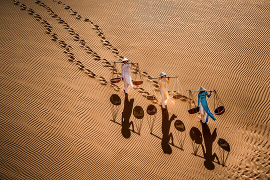 Aerial view of three Vietnamese ladies in traditional ao dai costumes, carrying straw baskets, traversing the sand dunes, their shadows stretching long, Mui Ne, Phan Thiet, Vietnam.