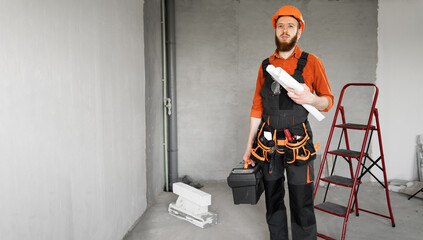 Bearded builder in hardhat holding toolbox and blueprints in apartment before renovation.