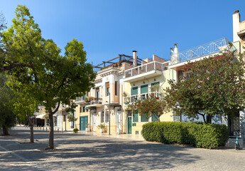 Quiet Athens neighborhood pedestrian street with pastel homes and balconies, trees and hedges, blue...