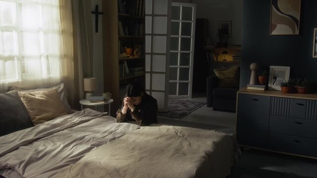 Brunette young woman praying with her head bowed and hands clasped while leaning on bed with elbows in cozy apartment