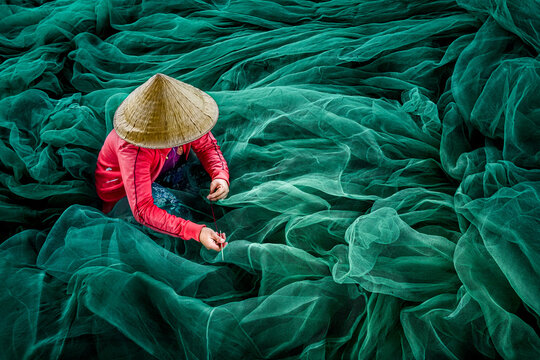 Aerial view of a woman in a conical hat mending a sea of vibrant turquoise fishing nets in sharp contrast to her bright pink top, Phan Rang, Thap Cham, Vietnam.