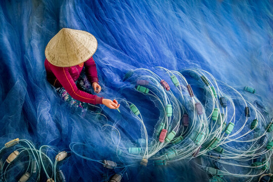 Aerial view of a person in a conical hat mending a vibrant blue fishing net, its intricate weave contrasting with the bright threads, Phan Rang, Thap Cham, Vietnam.