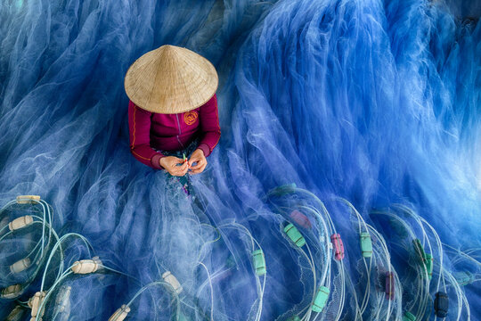 Aerial view of a person in a conical hat mending the vibrant blue fishing nets, creating a striking contrast of color and texture, Phan Rang, Thap Cham, Vietnam.