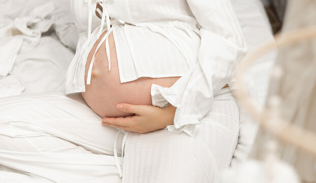 Pregnant woman in a bright nursery holding baby clothes near a crib. Soft natural light, cozy home interior, motherhood anticipation, maternity lifestyle, family love, neutral tones, calm atmosphere. - Powered by Adobe