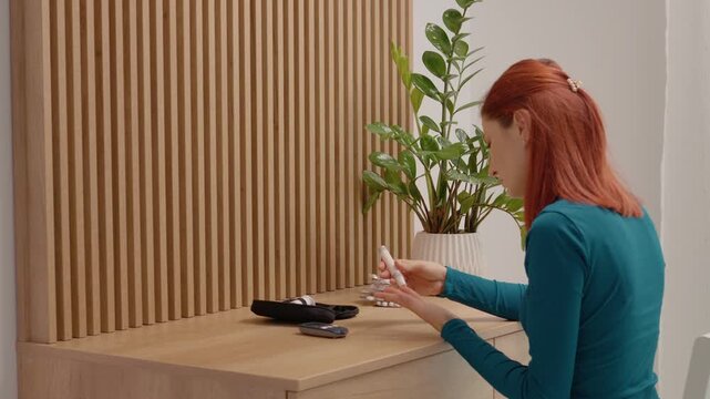 A woman tests her blood sugar by pricking her finger with a device. She sits at a table with a glucometer and a plant nearby, checking her glucose levels.