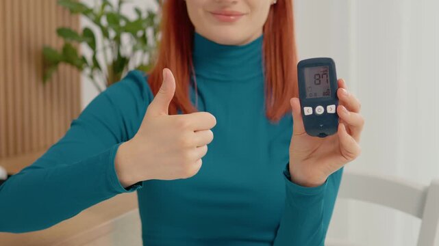 A woman displays a glucometer reading of 87 mg dL with a smile. She gives a thumbs up to show approval of her blood sugar level.