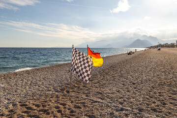 Chequered and lifeguard flags on a pebble beach against a mountain silhoutte and sea. Wide view of Konyaalti coast in Antalya, Turkey. Sunny autumn day in the Mediterranean resort. October, ripples.