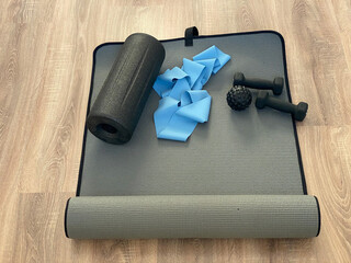 Fitness equipment arranged on a gray exercise mat, including a foam roller, resistance bands, dumbbells, and a massage ball on a wooden floor