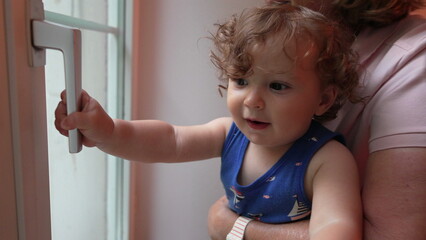 Curly-haired toddler held by grandmother reaches to touch door handle while looking outside with...