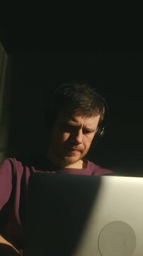 Young man wearing headphones concentrating on his laptop, his face illuminated by a single ray of sunlight in a dark room, creating a dramatic and pensive atmosphere of deep focus. Vertical shot.