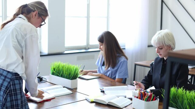 Three women collaborate at a sunlit desk, reviewing notebooks and discussing work in a bright, modern office or classroom setting.