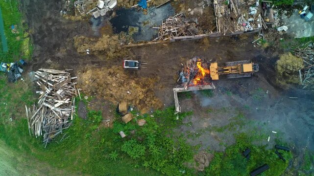 Aerial view descending downwards to a track loader containing a fire as people dismantle an old farm barn and burn it.