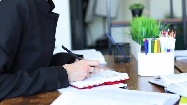 A person in a black blazer writes notes in a red notebook at a wooden desk, surrounded by books and colorful pens, focused on work or study.