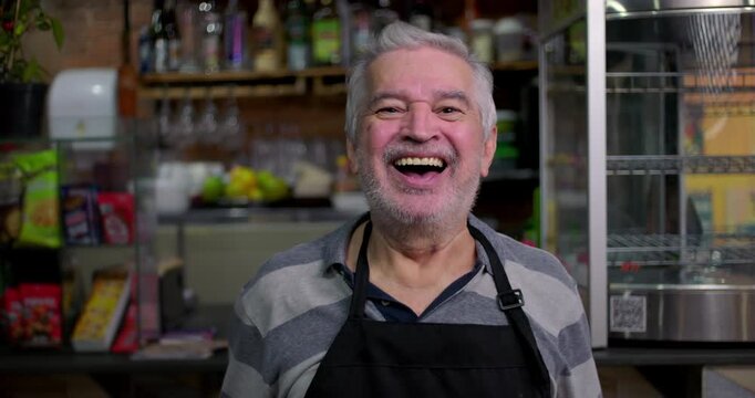 Older caf&eacute; owner laughing openly behind counter expressing joy friendliness approachability and human warmth within familiar rhythm of small neighborhood business