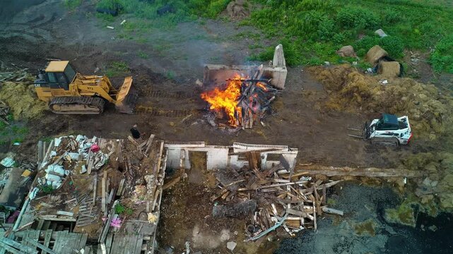An aerial shot coming down to a man adding planks of wood to a large fire as they dismantle and burn a barn.