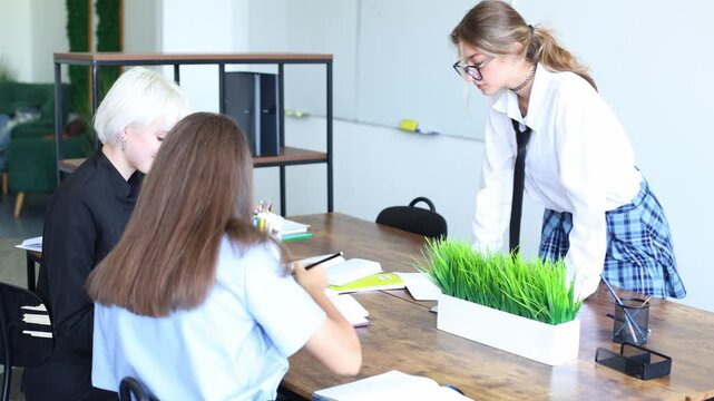A girls in a white shirt leans over a desk to discuss notes with two seated colleagues in a bright, modern office or classroom.