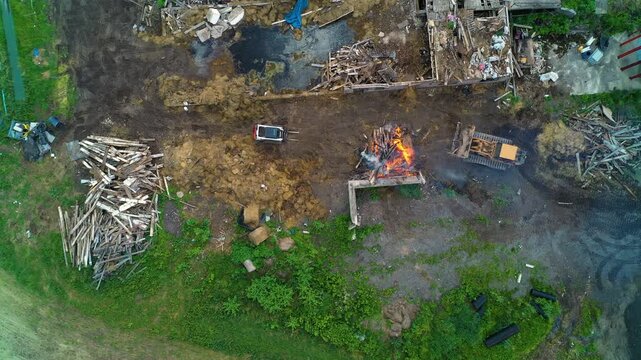 An aerial view of a rugged worksite with scattered lumber, debris piles, a track loader containing a fire, muddy ground and patches of green vegetation.