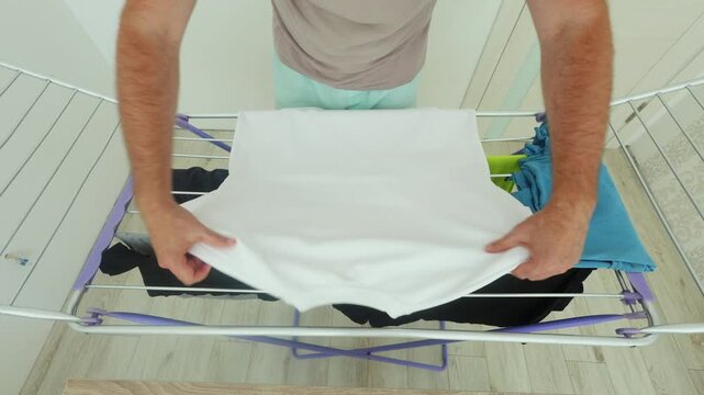 A man folds a white t-shirt neatly on a drying rack, demonstrating an efficient laundry folding technique in a tidy, modern home setting.