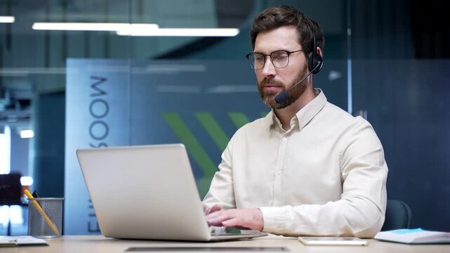 Male call center operator wearing headset working on laptop in modern office. Corporate customer support agent talking to client on helpline, providing service assistance, telesales, technical support