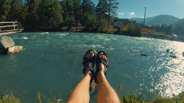 First-person perspective of a man relaxing, feet dangling over a turbulent turquoise river. Scenic mountain landscape with forests under a clear blue sky. Evokes freedom, adventure, and travel