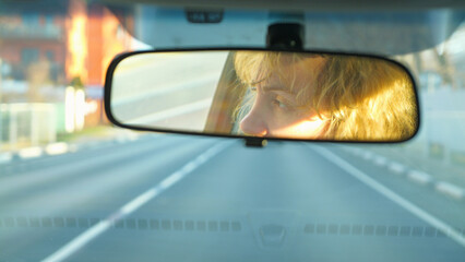 A woman's face reflects in a car's rearview mirror during a road trip. The scene, shot from a driver's POV, captures golden hour light, conveying a mood of travel and introspection