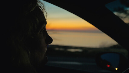 Pensive woman's silhouette in profile looking out a car window at a scenic coastal sunset. Moody, cinematic shot with warm colors a contemplative atmosphere. Concept of travel, journey, reflection