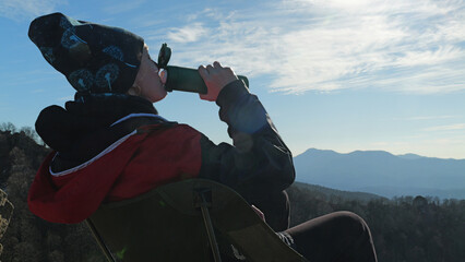 A woman in a windbreaker sits in a camp chair, resting after a hike. She drinks from a thermos and enjoys the picturesque view of the mountain range under a bright sunny sky. Relaxing in nature