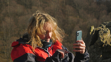 A young woman with windblown blonde hair in a red jacket is sitting outdoors and using her smartphone. The background shows a rocky, natural landscape. Natural sunlight
