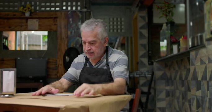 Older caf&eacute; owner works through handwritten calculations at table conveying persistence worry and emotional weight of trying to close month finances during difficult period