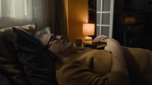 Mature man with glasses putting bible book on his chest and closing eyes while lying on bed with cushions under head