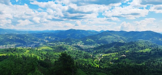 Panoramic mountain valley with lush green forests, meadows and winding road under blue sky with clouds, summer hiking landscape, scenic nature panorama

