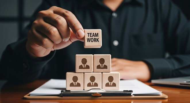 A person's hand places a 'TEAM WORK' block on a pyramid of human icon blocks, symbolizing collaboration and building a strong team in a business setting.