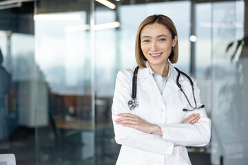 Young female doctor in a lab coat with stethoscope stands confidently with arms crossed, smiling warmly in a bright modern clinic, conveying professionalism and care © Liubomir