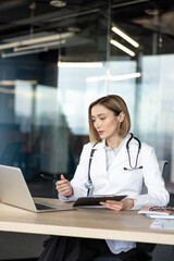 Fototapeta premium Female doctor in a white coat and stethoscope sitting at a desk, holding a pen and a tablet, pointing at a laptop screen during a virtual medical consultation or online conference