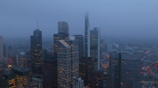 Aerial view of Frankfurt's financial district at dusk, featuring illuminated skyscrapers and office buildings partially obscured by dense fog