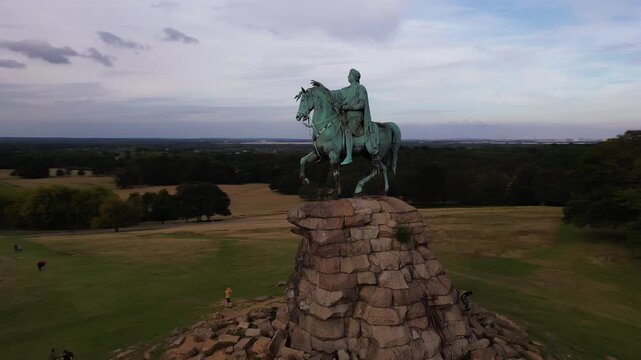 An equestrian statue stands atop a rocky monument. Below, rolling hills and trees stretch to the horizon under a cloudy sky.