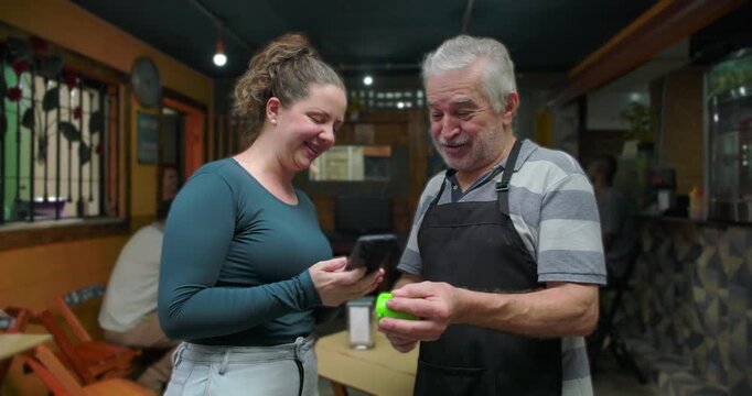 Small business caf&eacute; owner processing contactless payment with young woman inside neighborhood restaurant expressing friendly service trust cooperation modern customer experience