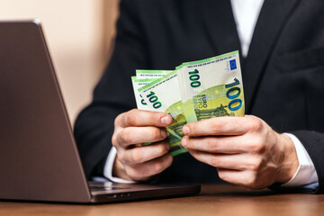 Businessman in a black suit holding several 100 euro banknotes while sitting at a desk with a laptop, indicating financial transactions or investments